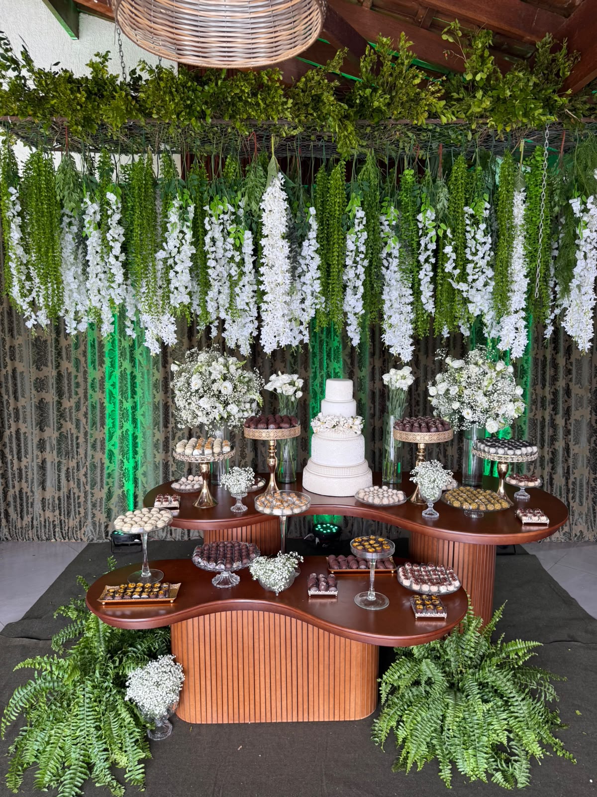 Mesa de casamento com bolo branco e doces variados sob flores brancas